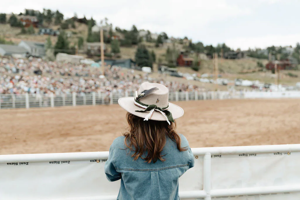 The kind of western polish that belongs at Boots & Bubbles. Person in a denim jacket wearing a custom cream western hat while watching a rodeo arena
