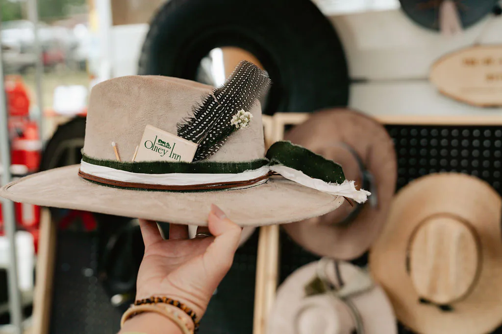 Start with a hat shape, then add your own finish. Hands holding a cream custom hat with feather, card, and band details at the hat bar