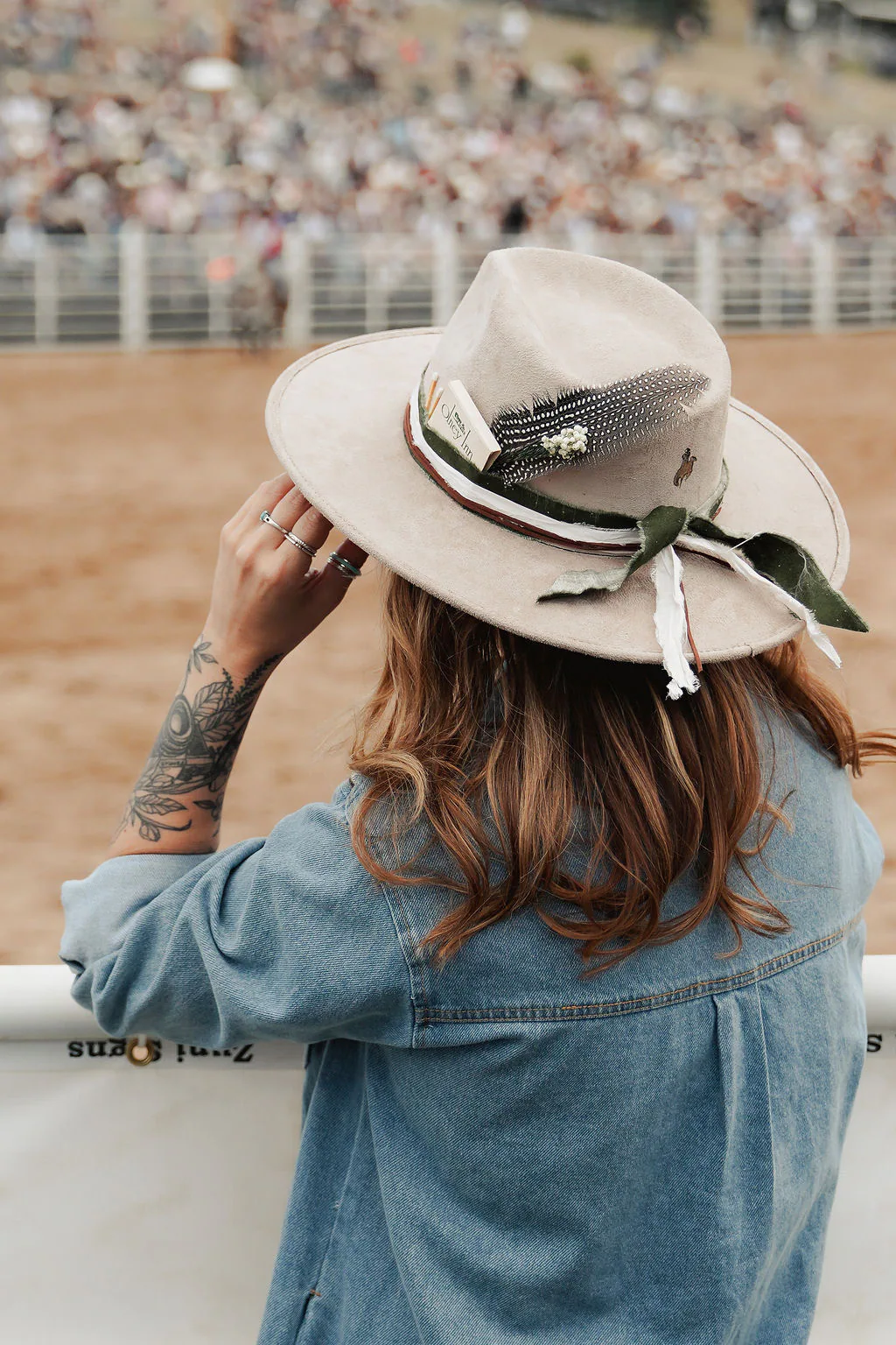 A finished western hat styled with ribbon and feather details. Person at an arena rail wearing a cream western hat with feather and ribbon details