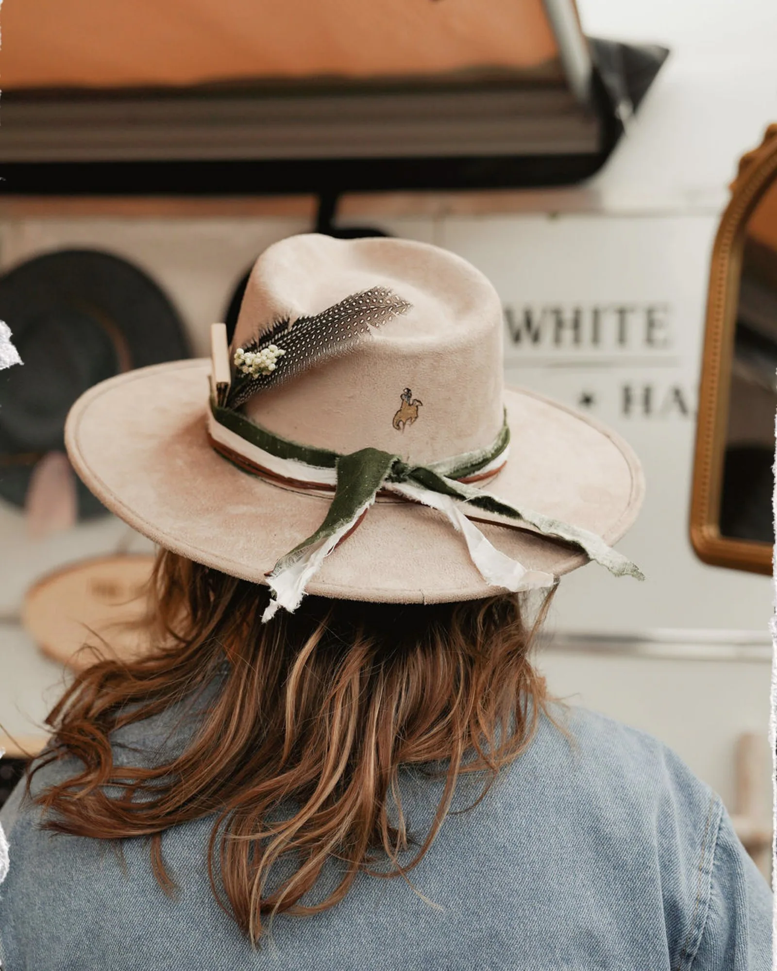 Custom details, texture, bands, feathers, and a western finish. Finished cream western hat with a ribbon band and feather accent on display